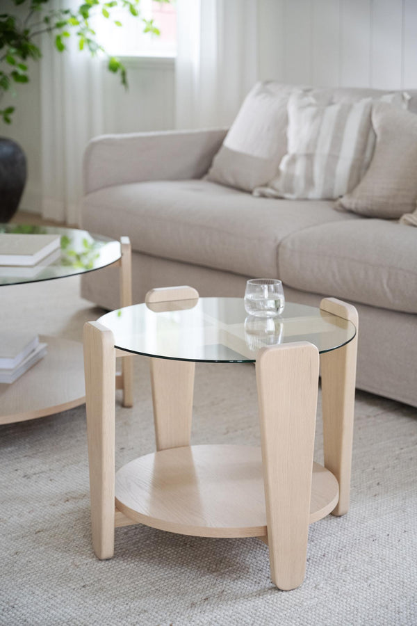 Runt soffbord i ljust trä med glaskiva och hylla, ställt framför beige soffa i modernt vardagsrum