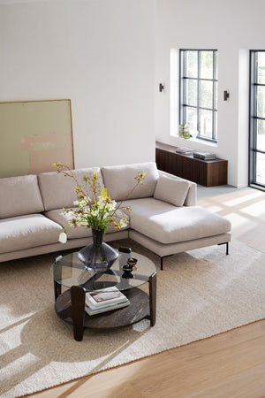 Ljust vardagsrum med beige hörnsoffa, runt soffbord i glas och trä, samt konstram mot vit vägg