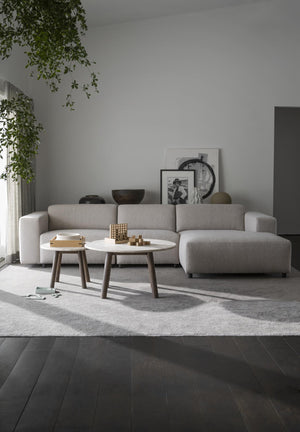 Minimalistisk ljusgrå soffa med schäslong och två runda soffbord i vardagsrum med mörkt trägolv