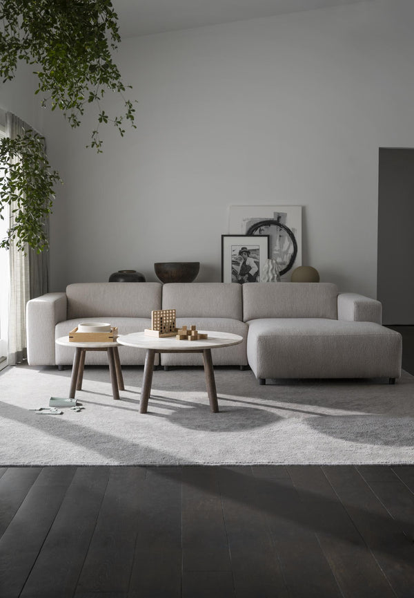 Minimalistisk ljusgrå soffa med schäslong och två runda soffbord i vardagsrum med mörkt trägolv