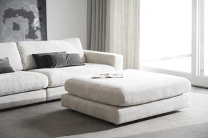 Vit modulsoffa med fotpall i vardagsrum med stora fönster och grå kuddar