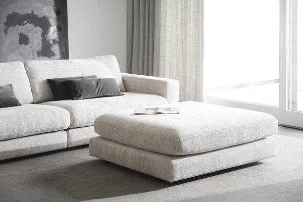 Vit modulsoffa med fotpall i vardagsrum med stora fönster och grå kuddar