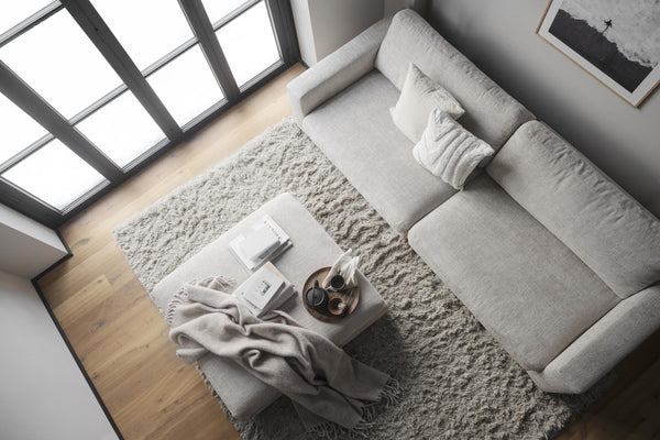 Grå modern soffa med kuddar och pläd, vit shaggy matta och soffbord i ljust vardagsrum med stora fönster