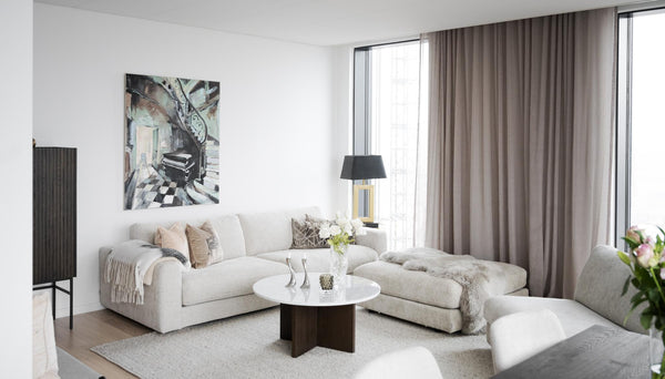 Modernt vardagsrum med beige hörnsoffa, runt soffbord, tavla och gråa gardiner vid stora fönster