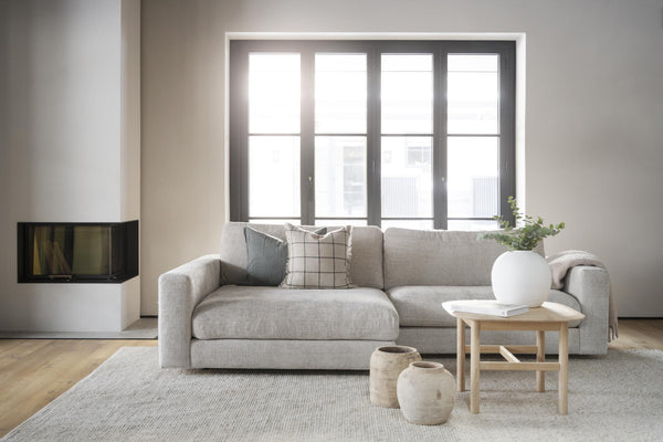 Ljust vardagsrum med grå soffa, soffbord i trä, vit vas med gröna växter och modern öppen spis