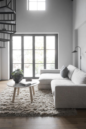 Minimalistiskt vardagsrum med grå soffa, rund soffbord och fluffig matta framför glasdörrar med naturligt ljus