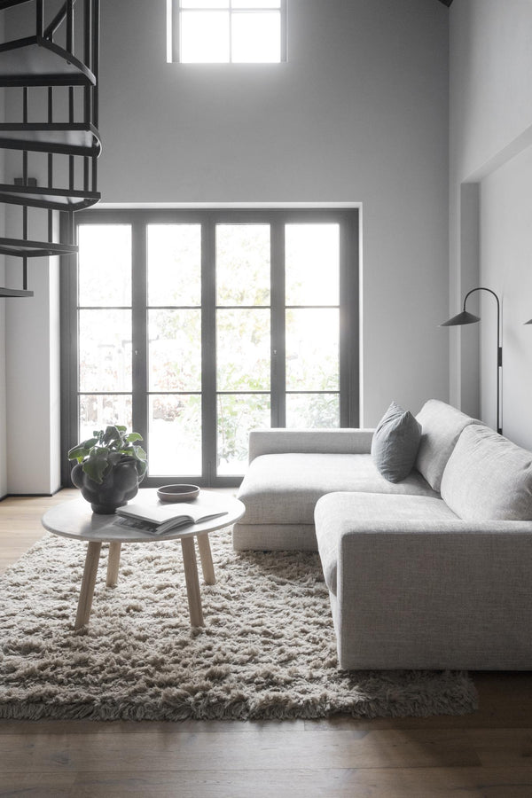 Minimalistiskt vardagsrum med grå soffa, rund soffbord och fluffig matta framför glasdörrar med naturligt ljus