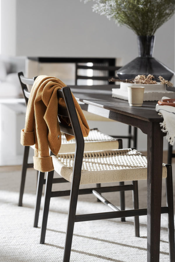 Matbord i mörkt trä med stol med flätad sits och senapsgul filt, kaffekopp och torkade blommor