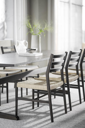 Matbord i grått trä med svarta stolar med flätade naturliga sittdynor i ljus matsal