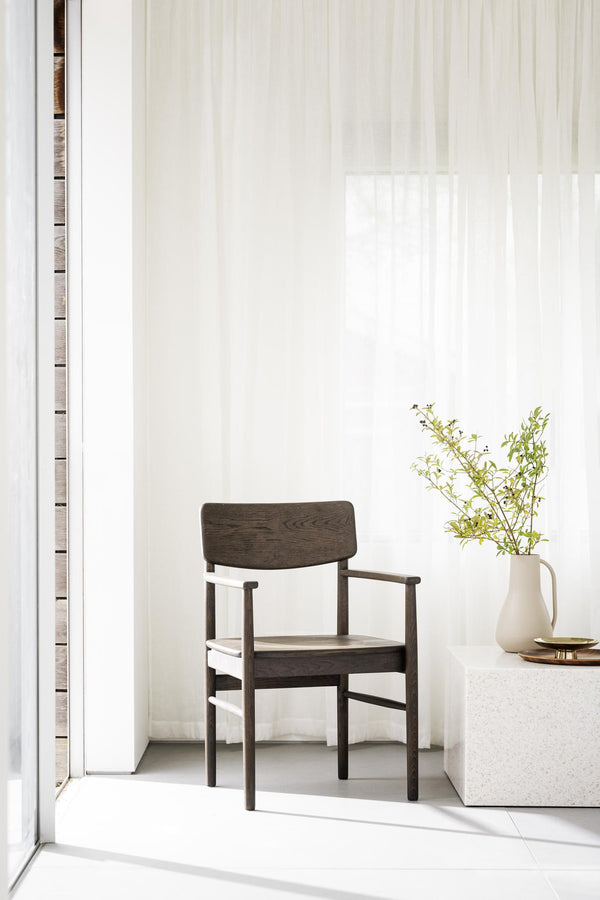 Mörkbrun träarmstol med minimalistisk design i ljust skandinaviskt rum med vit vas och gröna växter