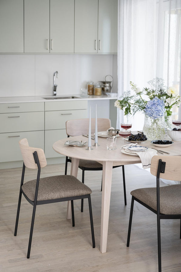 Ljust kök med runt matbord i trä och stolar med grå dynor, dekorerat med blommor och ljusstakar