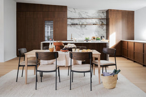 Modern matplats med ljust träbord, stolar med beige dynor och mörka träskåp i köket