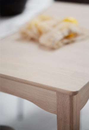 Närbild på hörn av matbord i ljust trä med tallrik med mat i oskärpa på bordsskivan