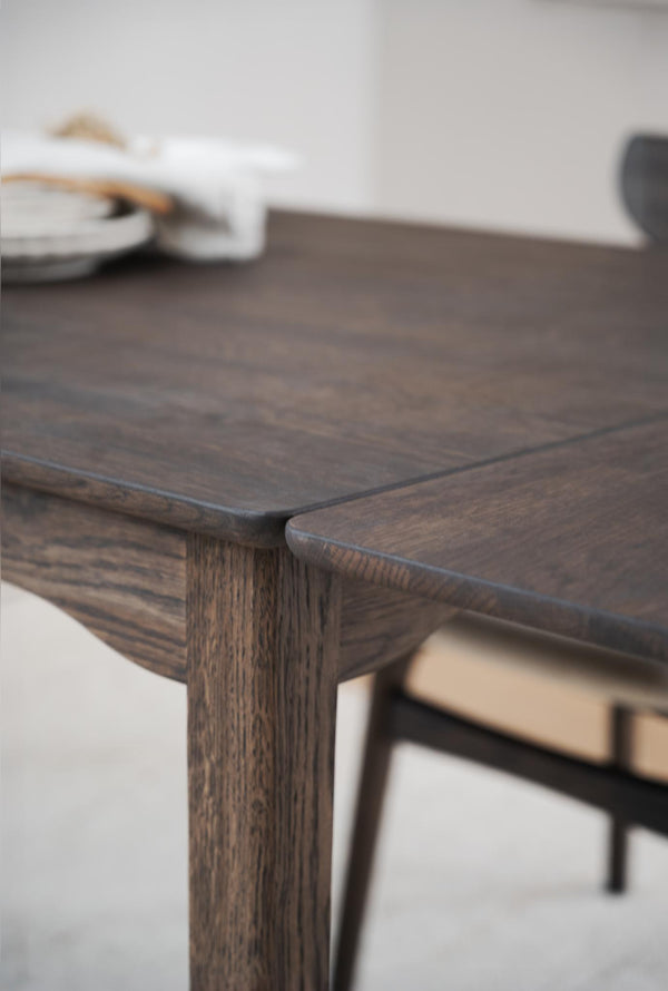 Närbildsdetalj av mörkbrunt träbord med synlig trästruktur och runda hörn, stol i bakgrunden