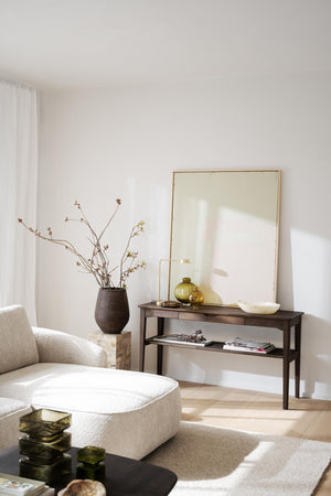 Minimalistiskt vardagsrum med beige sittpuff, mörkt konsolbord, stor guldspegel och dekorativ vas med kvistar