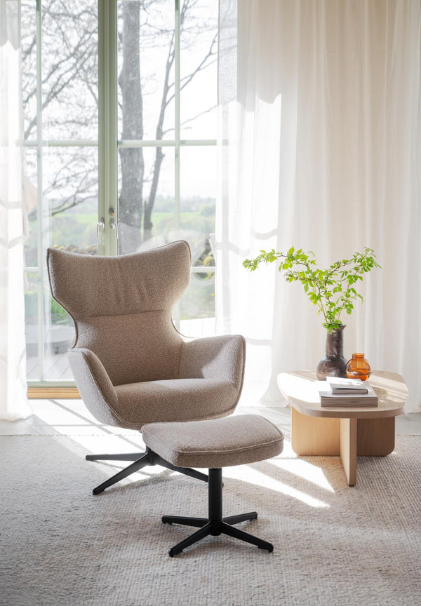 Beige fåtölj med fotpall och svart stjärnfot framför fönster med runt soffbord och växtdekoration