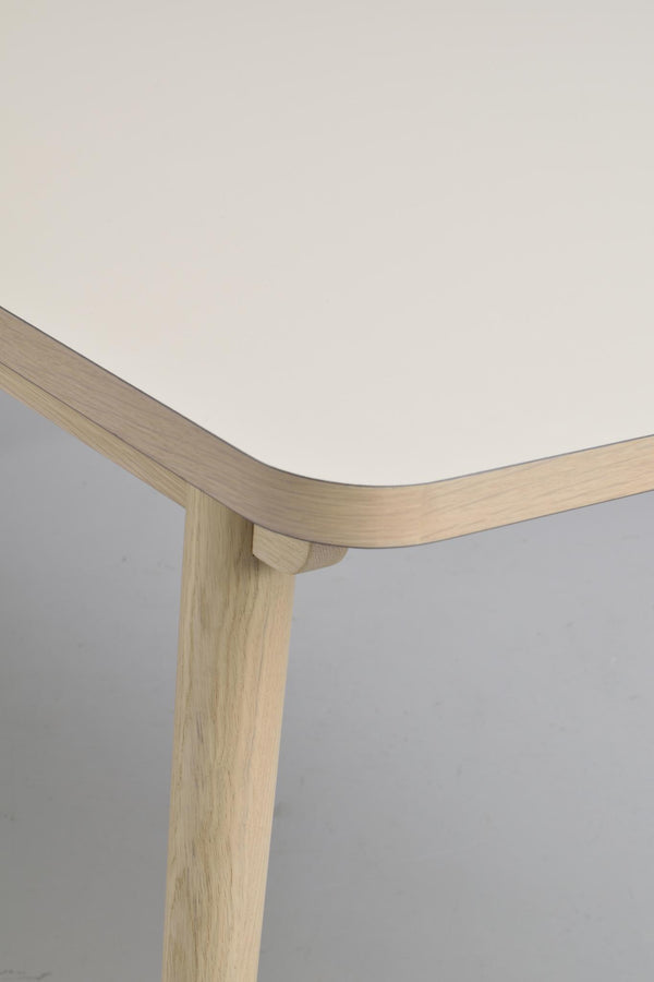 Närbild av bordshörn med vit laminatskiva och träkant i ljus ek, samt träben i modern skandinavisk design