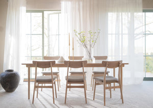 Matbord i ek med sex stolar i ljust trä runt ett vackert uppdukat bord med vita gardiner i bakgrunden