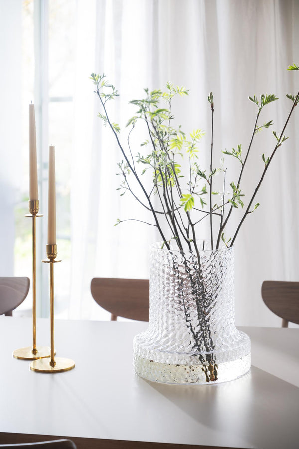 Glasklar vas med struktur innehåller gröna kvistar på matbord bredvid gyllene ljusstakar