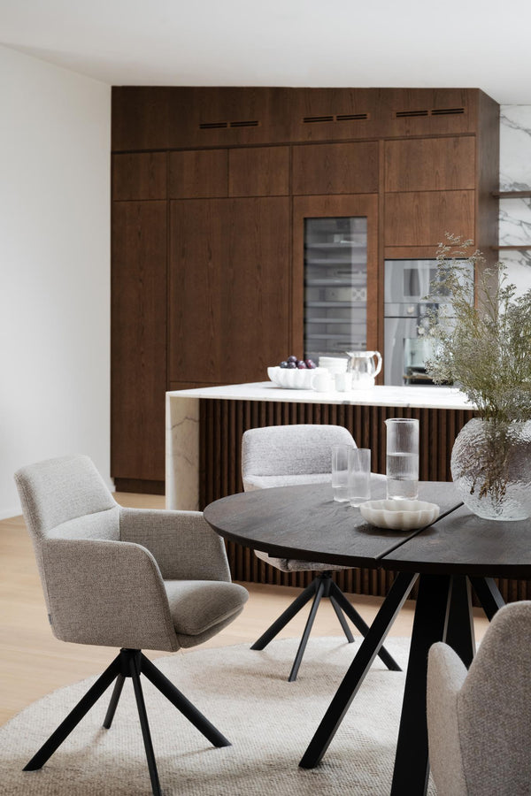 Matgrupp med svartbetsade stolar i grå klädsel och svart runt matbord mot mörkt träkök med vit bänkskiva