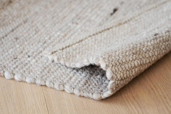 Beige handvävd matta i ull med texturerat mönster, uppvikt hörn visar baksidan mot ljust trägolv