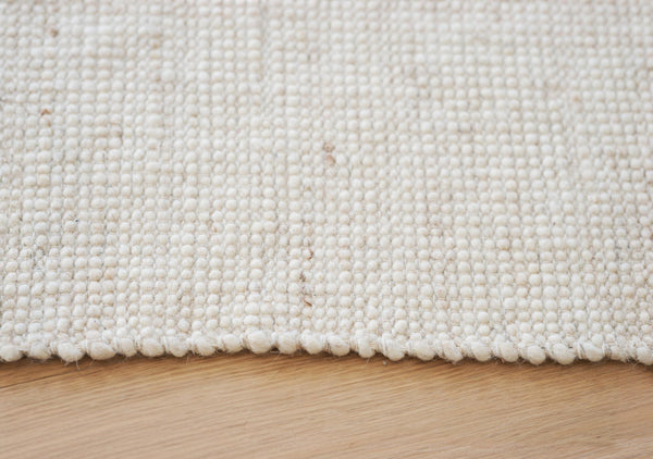 Närbild på vit/beige handvävd ull-matta med texturerad yta mot ljust trägolv