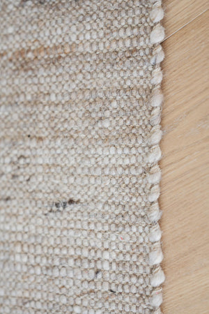 Närbild av beige matta med flätad kant mot ljust trägolv, visar texturen och detaljerna i vävningen