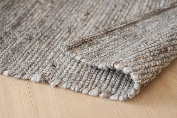 Närbild på handvävd ullmatta i beige och grå toner med synlig textur och fransad kant på trägolv