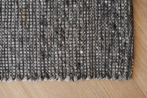 Närbild på grå handvävd ullmatta med strukturerad yta och synlig fransad kant på trägolv