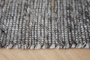 Närbild på handvävd matta i grå och beige nyanser med fransad kant mot trägolv