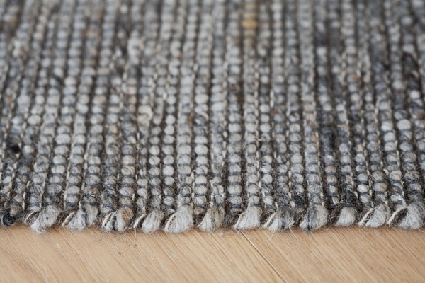 Närbild på handvävd matta i grå och beige nyanser med fransad kant mot trägolv