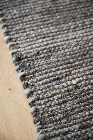 Närbild av handvävd ullmatta i gråa toner på ljust trägolv med synlig struktur och kant