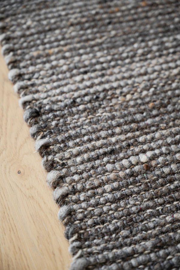 Närbild av handvävd ullmatta i gråa toner på ljust trägolv med synlig struktur och kant