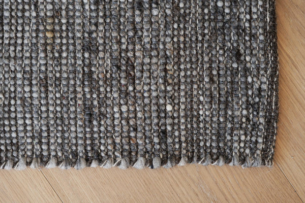Närbild på gråmelerad vävd matta med strutna fransar mot ljust trägolv