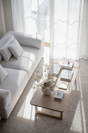 Ljust vardagsrum med grå soffa, vitt soffbord och genomskinliga gardiner med solljus