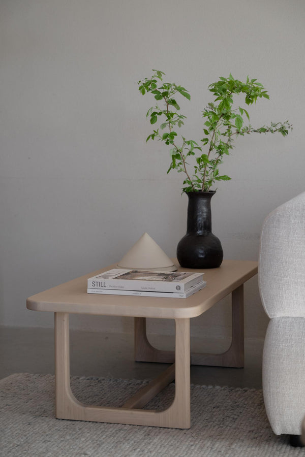 Soffbord i ljust trä med böcker, svart vas med gröna kvistar och dekorativ vit pyramid bredvid grå fåtölj