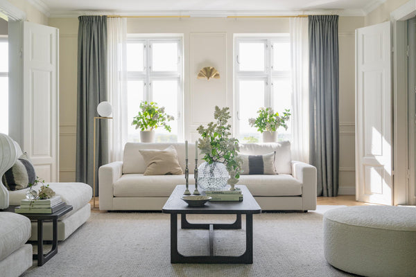 Ljust vardagsrum med beige soffor, gråa gardiner, svartt soffbord och stora fönster med växter