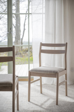 Ljus trästol med stoppad sits vid fönster med vita gardiner och utsikt mot trädgård