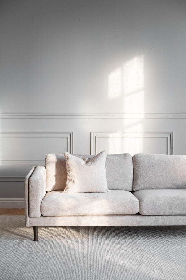 Ljusgrå soffa med kuddar mot vit vägg med spegelpaneler och fönsterskugga