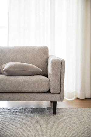 Beige tvåsitssoffa med kudde och svarta ben på ljus matta framför vit gardin