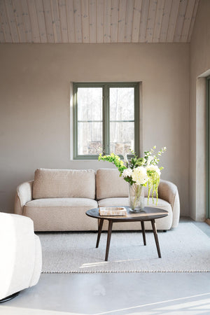 Beige tvåsitssoffa med rundt soffbord i vardagsrum med minimalistisk inredning och grönt fönster
