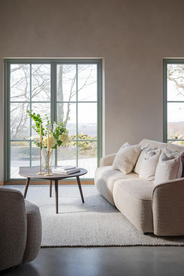 Ljust vardagsrum med beige bouclésofa, runt soffbord och fönster med utsikt över sjö