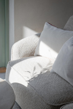 Vit kudde med röd kantband på grå fåtölj med cremefärgad pläd i solljus