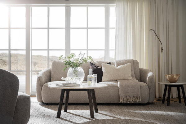 Beige tvåsitsarsoffa med ljusa kuddar och pläd framför stora fönster i minimalistiskt vardagsrum