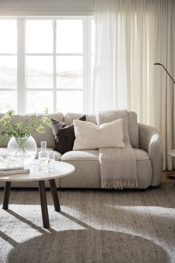 Beige bouclésofa med kuddar och pläd i ljust vardagsrum med runt soffbord och stora fönster