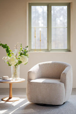 Beige fåtölj i teddyfleece med rund sits bredvid sidobord med vas och blommor vid fönster