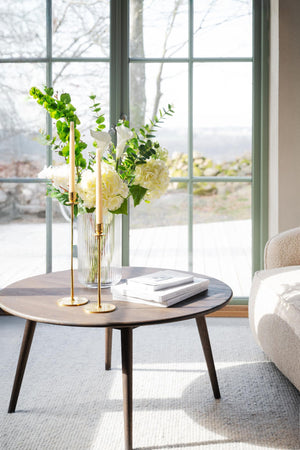 Runt soffbord i trä med glasvas, ljusstakar i mässing och böcker framför fönster i vardagsrum