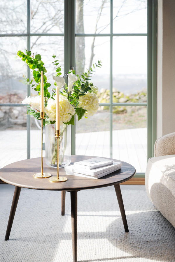 Runt soffbord i trä med glasvas, ljusstakar i mässing och böcker framför fönster i vardagsrum