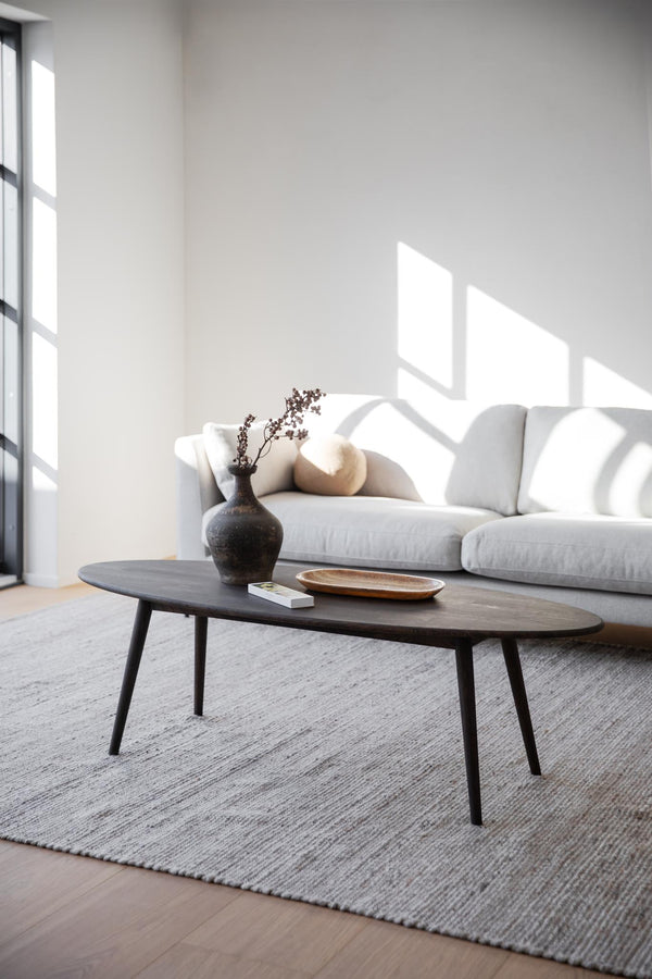 Ovalt soffbord i svart trä med spindelben i minimalistiskt vardagsrum med grå soffa och ljusinsläpp