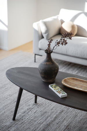 Mörkt ovalt soffbord i trä med keramikvas, träskål och dekorativa grenar i minimalistiskt vardagsrum